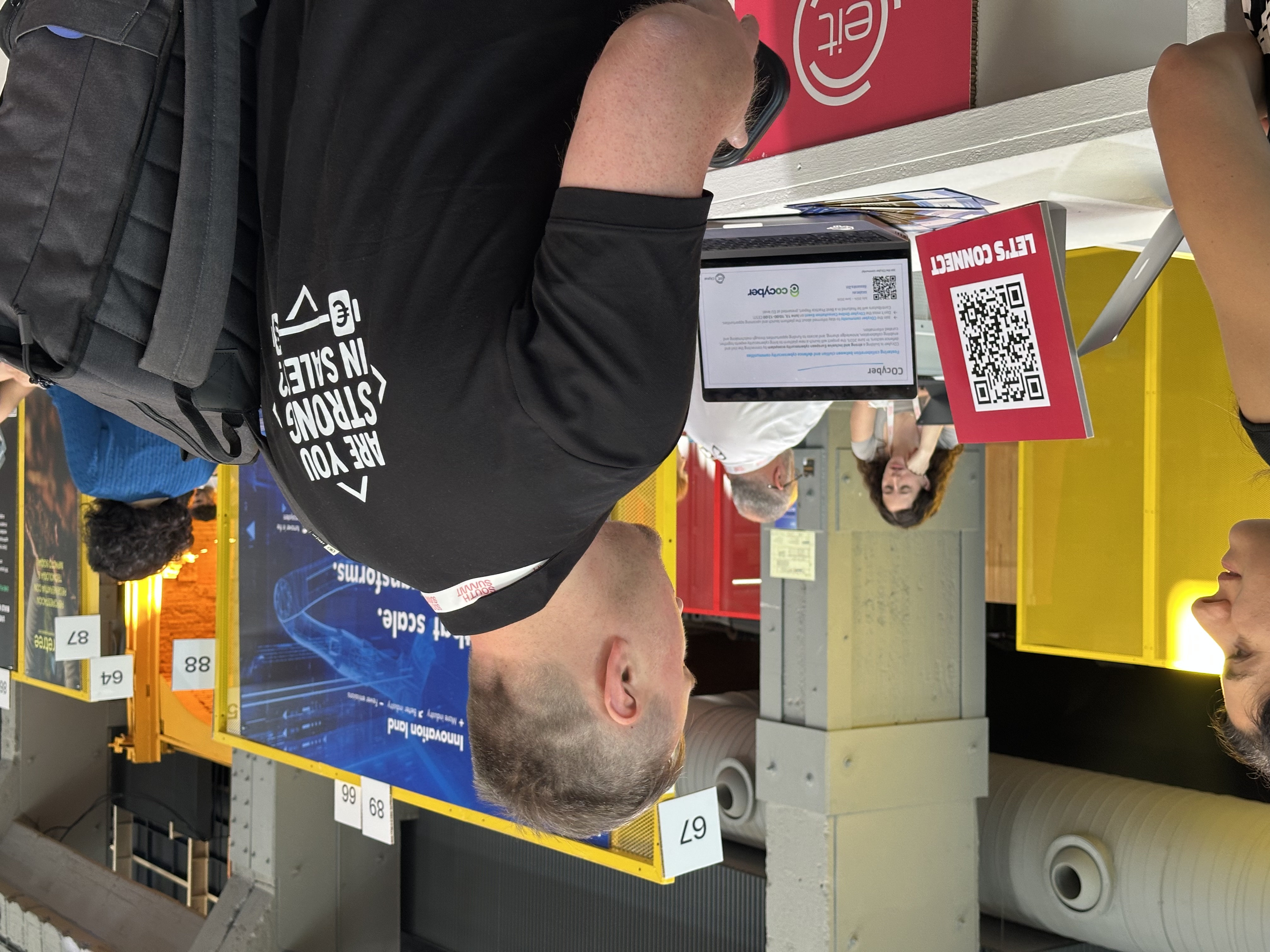 A COcyber slide is displayed on a laptop at the EIT Digital booth, surrounded by event banners and visitors.