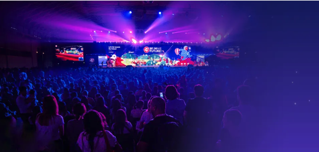 A brightly lit stage with "We Make Future" in bold letters, viewed by a crowd under purple and blue lighting.